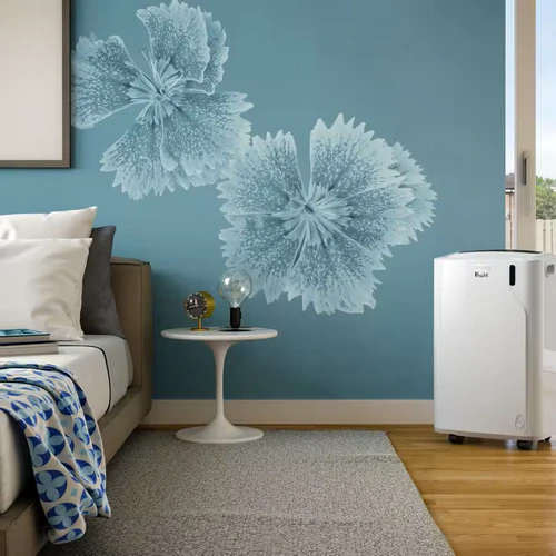 The compact De'Longhi Pinguino PAC EM82K.1 positioned on a wooden floor in a contemporary bedroom setup next to a bed, set against a blue wall with decorative floral artwork.